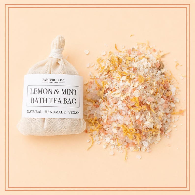 May include: A beige fabric bath tea bag with a white label reading PAMPEROLOGY LONDON LEMON & MINT BATH TEA BAG NATURAL HANDMADE VEGAN, positioned next to a loose pile of pink Himalayan salt, dried yellow flower petals, and white oat flakes.