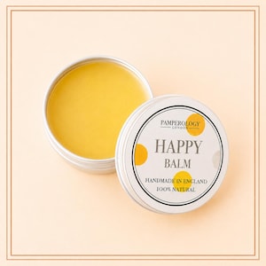 May include: A round silver metal tin containing yellow balm, with a white lid featuring yellow polka dots and black text that reads Pamperology London, Happy Balm, Handmade in England, and 100% Natural, displayed on a light peach background.