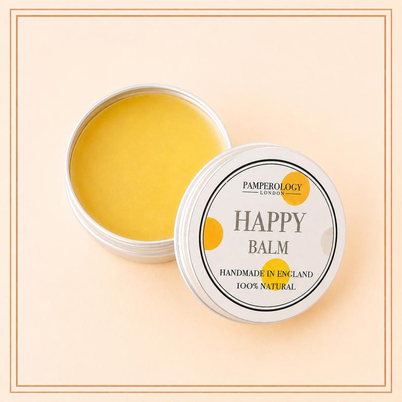 May include: A round silver metal tin containing yellow balm, with a white lid featuring yellow polka dots and black text that reads Pamperology London, Happy Balm, Handmade in England, and 100% Natural, displayed on a light peach background.