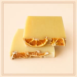 May include: Two rectangular bars of pale yellow soap stacked on top of each other. Each bar is embedded with dried orange slices along the bottom edge, featuring a textured, rustic appearance with visible citrus segments in shades of deep orange.