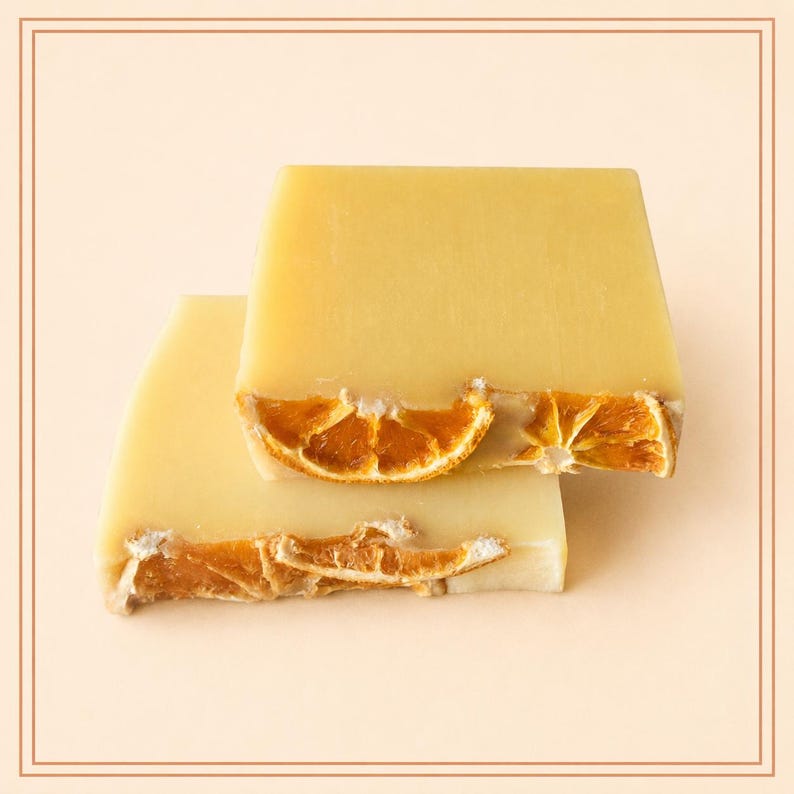 May include: Two rectangular bars of pale yellow soap stacked on top of each other. Each bar is embedded with dried orange slices along the bottom edge, featuring a textured, rustic appearance with visible citrus segments in shades of deep orange.