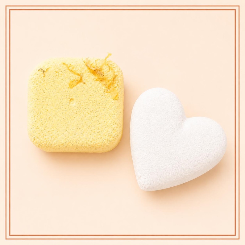 May include: A square yellow bath bomb with embedded dried flower petals and a smooth white heart-shaped bath bomb. Both items feature a porous, compressed texture and are positioned side by side against a solid light peach-colored background.