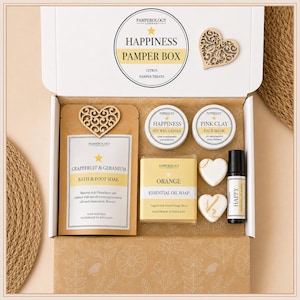 May include: A white gift box labelled Happiness Pamper Box containing a grapefruit and geranium bath soak, a lime and mandarin soy wax candle, a pink clay face mask, an orange essential oil soap, two heart-shaped items, and a small roller bottle.
