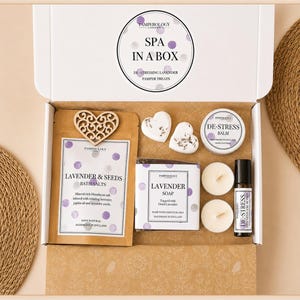 May include: A white gift box labelled SPA IN A BOX containing a pouch of Lavender & Seeds bath salts with a wooden heart, two heart-shaped soaps, a tin of De-Stress balm, two round tea light candles, and a bottle of De-Stress essential oil blend.