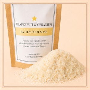 May include: A brown and white pouch labeled Grapefruit & Geranium Bath & Foot Soak, containing mineral rich Himalayan salt infused with essential oils and chamomile flowers, with a pile of coarse, light-colored salt crystals in the foreground.
