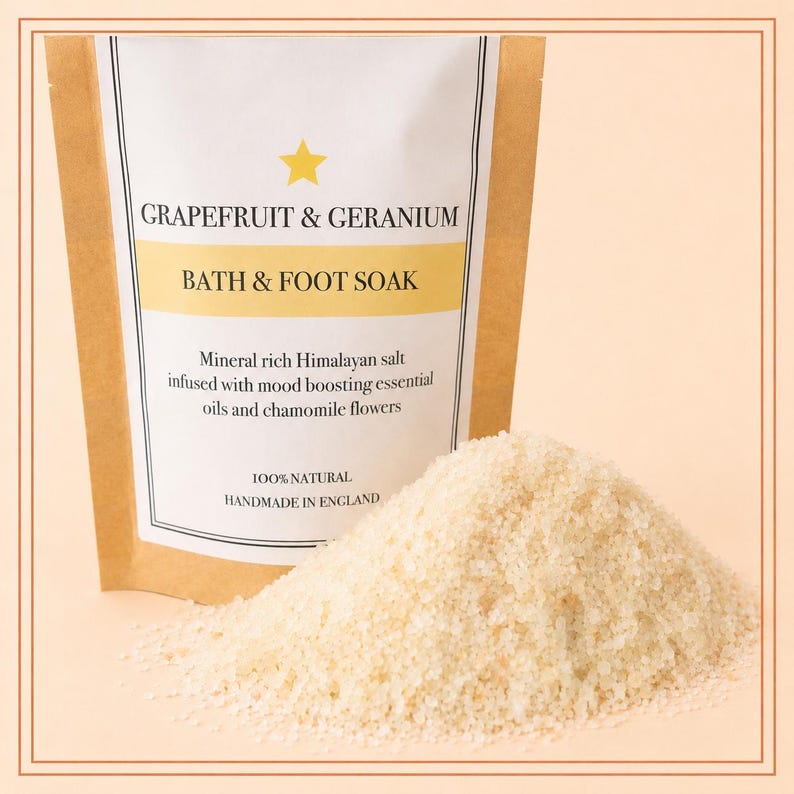 May include: A brown and white pouch labeled Grapefruit & Geranium Bath & Foot Soak, containing mineral rich Himalayan salt infused with essential oils and chamomile flowers, with a pile of coarse, light-colored salt crystals in the foreground.