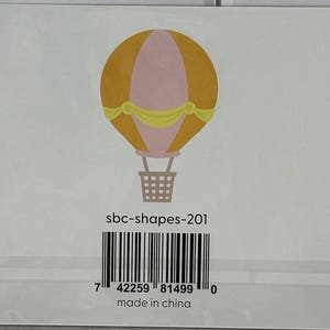 May include: A card featuring a hot air balloon illustration. The balloon is primarily mustard yellow with a pink center and a yellow ribbon. The basket is brown. The card has the text "sbc-shapes-201" and a barcode. The text "made in china" is also present.