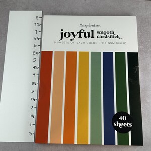 May include: A package of "joyful" smooth cardstock with 40 sheets. The cover features vertical stripes in various colors, including orange, yellow, green, and blue. The package is next to a white ruler with inch measurements.