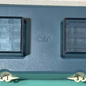 May include: A teal-coloured, rectangular tool with two square, transparent compartments. The tool has a textured surface and a logo in the centre. Numbered dials are on the bottom. The compartments have different internal patterns.