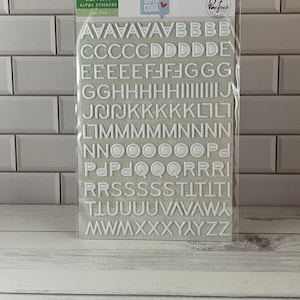 May include: A package of white puffy alphabet stickers. The stickers are in various capital letters and numbers. The package has the text "MINI PUFFY ALPHA STICKERS" and "SUPER COOL" on it. The background is a light green color.