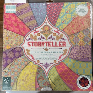 May include: A premium paper pad titled "Storyteller" displays a range of colourful paisley and floral patterns. The pad measures 12 x 12 inches (30.5 cm x 30.5 cm) and includes gem, pearlescent, and glitter effects. The paper is double-sided.