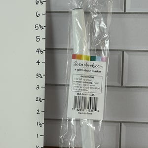 May include: A clear plastic package containing a white glitter brush marker. The marker has a white barrel and a black tip. The packaging includes a label with instructions and the text "Scrapbook.com glitter brush marker."