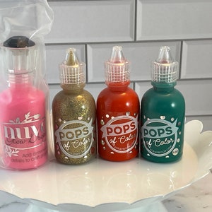 May include: A collection of craft supplies, including four bottles of "POPS of Color" in gold glitter, red, and green, and a pink "nuvo Glow Drops" bottle. The bottles are displayed on a white, scalloped-edge plate.
