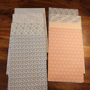 Op de afbeelding: Stapels bedrukte stoffen in verschillende ontwerpen en kleuren. De patronen omvatten bladeren, strepen en geometrische vormen in tinten blauw, groen, oranje en wit. De stof is netjes op een houten oppervlak gestapeld.