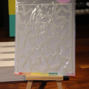 May include: A stencil sheet featuring a butterfly design, packaged in clear plastic. The package includes the text "Scrapbook.com" and "Butterflies". The stencil is displayed on a small wooden easel.