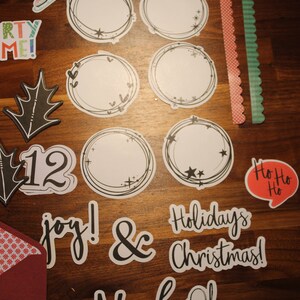 May include: A collection of Christmas-themed paper cutouts and stickers on a wooden surface. Includes circular frames, holiday greetings, and decorative elements like skates, holly, and tickets. The color palette features red, green, black, and white.