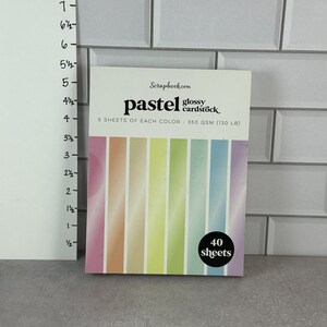 May include: A package of pastel glossy cardstock from Scrapbook.com. The cardstock features a gradient of colors, including pink, orange, yellow, green, blue, and purple. The package contains 40 sheets of 350 GSM (130 LB) cardstock.