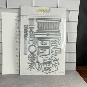 Pode incluir: Um pacote de matrizes de corte Honey Bee Stamps para materiais de artesanato em papel. Os cortes de matrizes prateados incluem formas de móveis, uma roda e outros elementos decorativos. O pacote está em uma superfície branca e cinza.