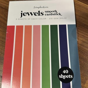 May include: A box of "Jewels" smooth cardstock from Scrapbook.com. The box features a colorful striped design with red, orange, pink, green, navy, and blue vertical stripes. It contains 40 sheets of 85lb paper, with 5 sheets of each color.