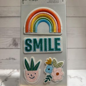 May include: A pack of four adhesive patches featuring a rainbow, the word "SMILE", a potted plant with a smiley face, and a cluster of flowers. The patches are on a white background with the text "pebbles" and "adhesive patches" printed on the top.