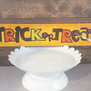 Könnte beinhalten: Ein Halloween-Banner mit den Worten "TRICK or TREAT" in orangefarbenen, gelben und lila Glitzerbuchstaben. Das Banner ist vor einem gelben Hintergrund platziert und wird über einem weißen Kuchenständer mit gewelltem Rand angezeigt.