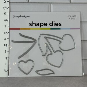May include: A package of shape dies from Scrapbook.com. The package contains several metal dies in the shape of cherries, leaves, and a heart. The package is white with the words "shape dies" in black and "cherries 4 pcs" in the upper right corner.