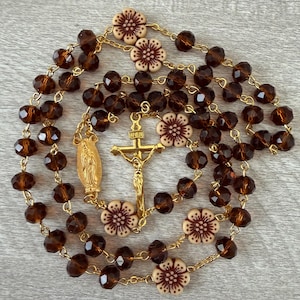 May include: A rosary featuring dark brown faceted glass beads and cream-colored flower-shaped beads with brown centers. The rosary includes a gold-toned metal crucifix and a gold-toned oval medal depicting a figure, all connected by gold-toned metal links.