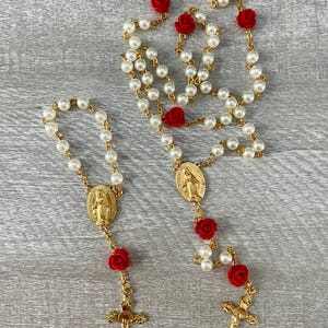 Pode incluir: Dois rosários com contas de pérolas falsas, detalhes de rosas vermelhas e componentes metálicos dourados. Um é uma pulseira, o outro um colar com um pingente de crucifixo e um pingente oval com uma figura religiosa.