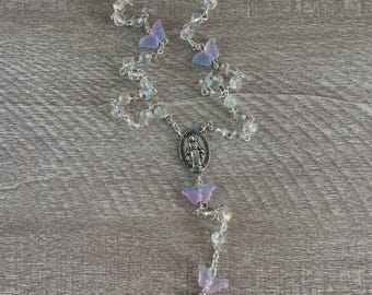 May include: A rosary with clear, iridescent beads and delicate butterfly accents in shades of lavender and pale blue. The rosary features a silver-toned crucifix and a central medallion with a religious figure.