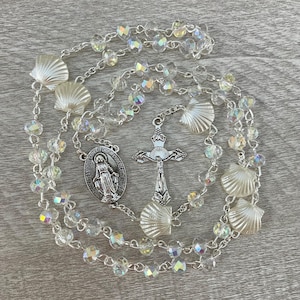 May include: A rosary with clear, iridescent beads and seashell accents. The silver-toned crucifix and Virgin Mary pendant are connected by a delicate chain. The rosary is laid out on a light gray wooden surface.