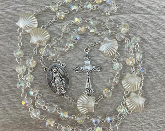 May include: A rosary with clear, iridescent beads and seashell accents. The silver-toned crucifix and Virgin Mary pendant are connected by a delicate chain. The rosary is laid out on a light gray wooden surface.