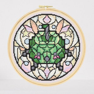Peut inclure: Un motif de broderie au point de croix circulaire représentant une créature géométrique verte avec des accents roses, entourée d'un motif coloré ressemblant à un vitrail.