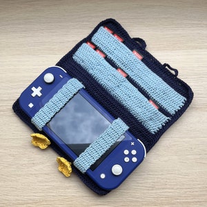 以下が含まれることがあります： 紺色の編み物ケースに、青色のNintendo Switch Liteコンソールが収められています。水色の編み物ストラップがコンソールを固定しています。2つの水色の編み物カートリッジホルダーが見えます。2つの小さな黄色の編み物アクセサリーも存在します。