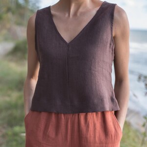 May include: A brown linen sleeveless top with a v-neckline paired with rust-coloured linen trousers. The model is standing outdoors with green grass and a blurred background.