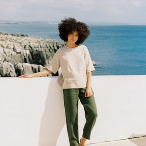 May include: A woman wearing a beige short-sleeved top and olive green pants. The outfit is paired with light brown sandals. The background features a white wall, the ocean, and a cloudy sky. The clothing appears to be made of linen.