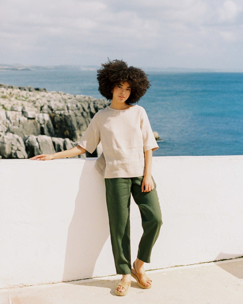 May include: A woman wearing a beige short-sleeved top and olive green pants. The outfit is paired with light brown sandals. The background features a white wall, the ocean, and a cloudy sky. The clothing appears to be made of linen.