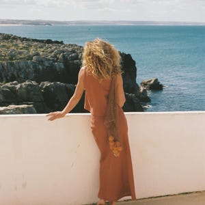 May include: A woman in a rust-colored sleeveless top and matching skirt stands on a white ledge overlooking the ocean. She holds a net bag filled with oranges. The sky is blue with scattered clouds, and the sea is a deep blue.