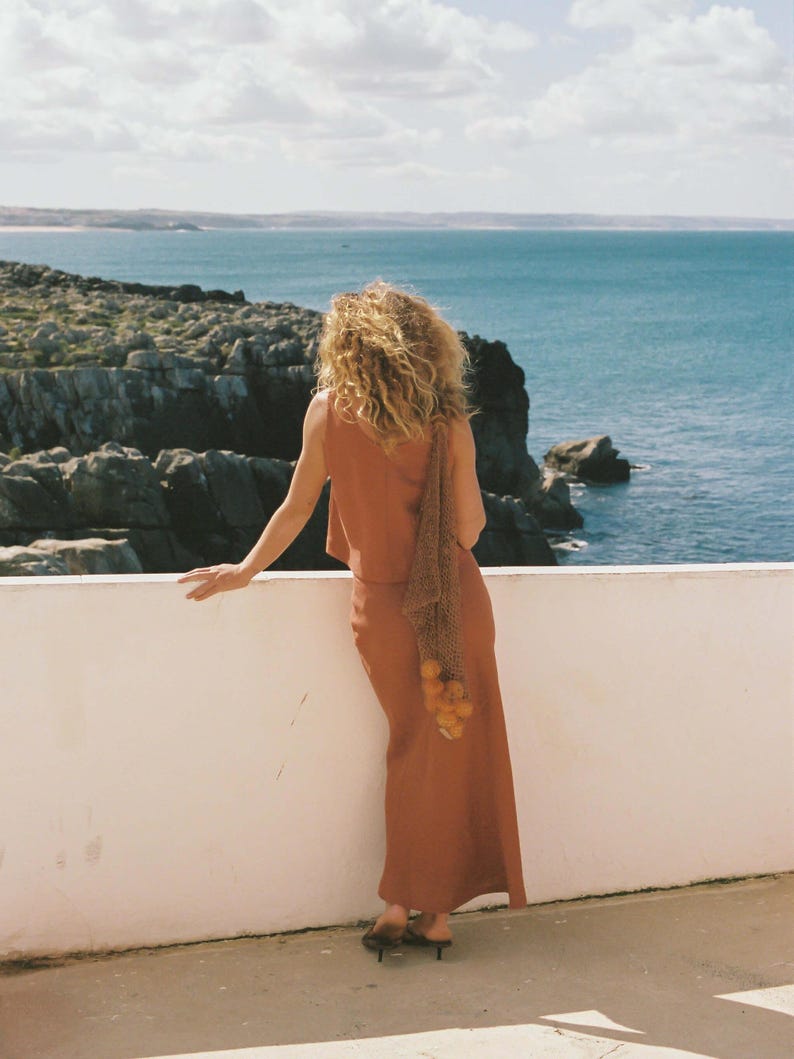 May include: A woman in a rust-colored sleeveless top and matching skirt stands on a white ledge overlooking the ocean. She holds a net bag filled with oranges. The sky is blue with scattered clouds, and the sea is a deep blue.