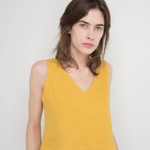 May include: A woman wearing a sleeveless, yellow, cropped top with a v-neck and black pants.