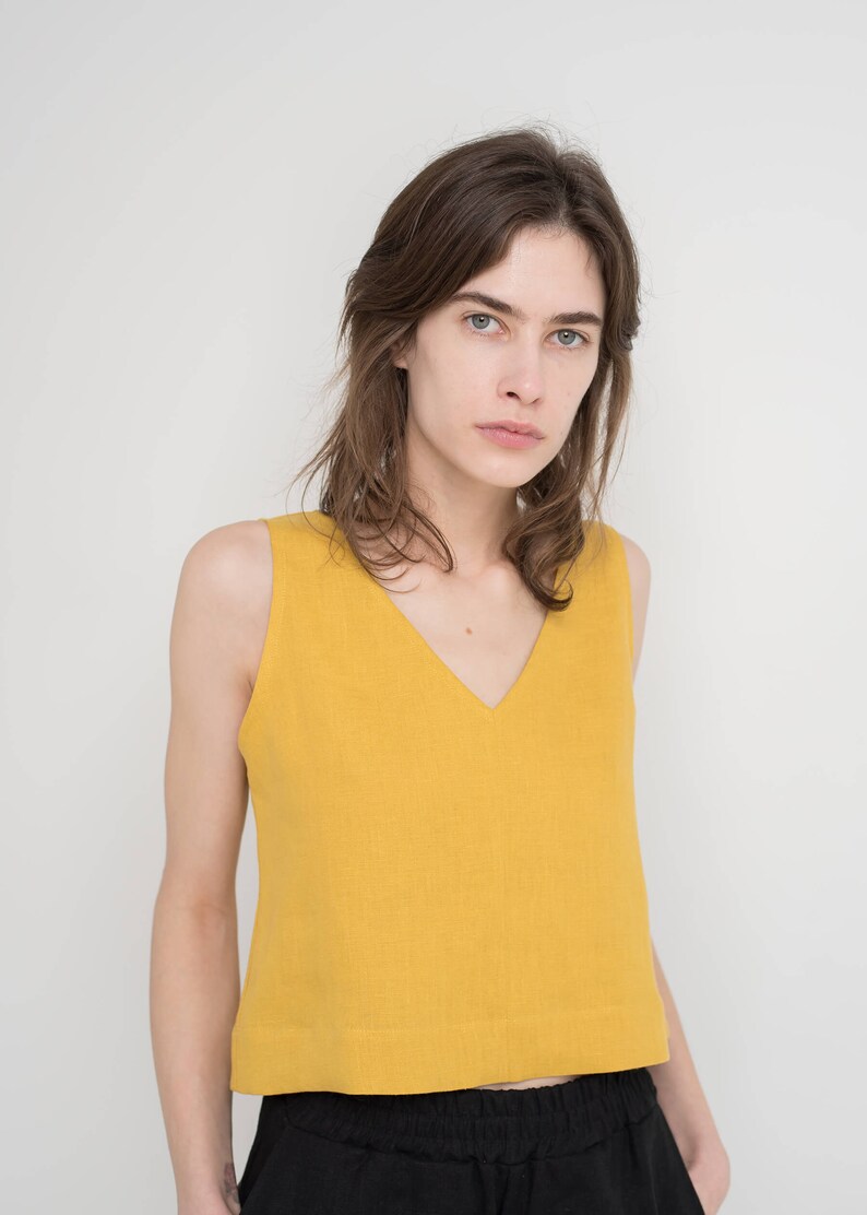 May include: A woman wearing a sleeveless, yellow, cropped top with a v-neck and black pants.