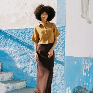 May include: A woman wearing a mustard-colored short-sleeved button-down shirt and a long, dark brown skirt. She is standing on a set of white concrete steps with a blue and white painted wall in the background. She is wearing sandals.