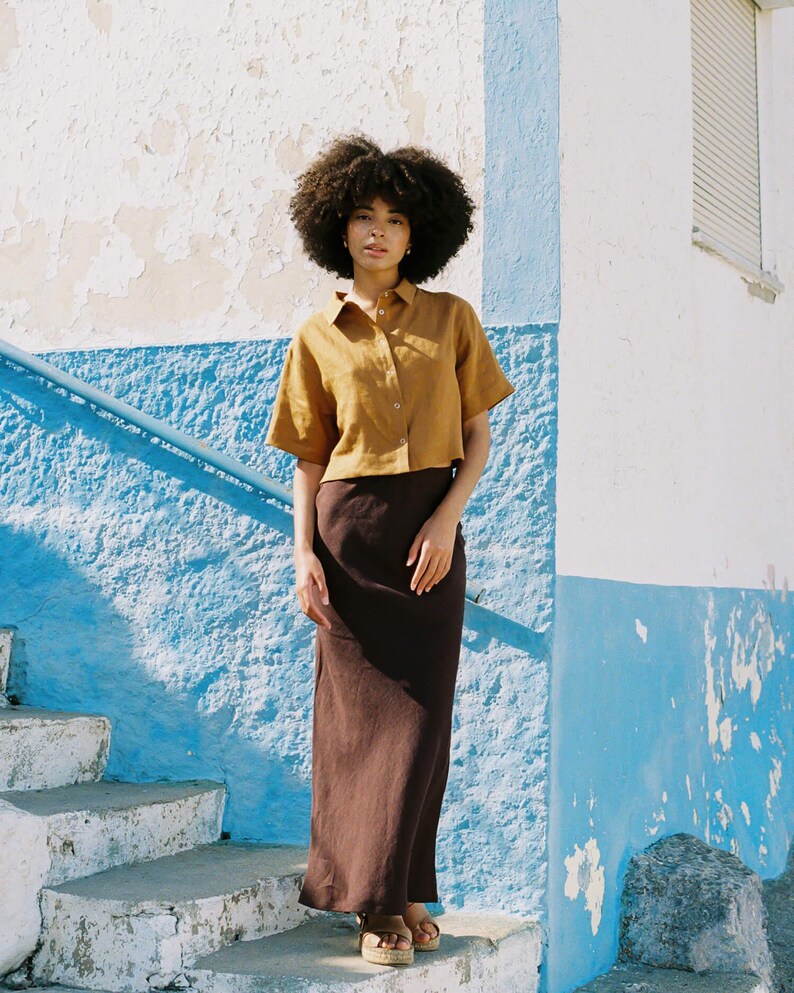 May include: A woman wearing a mustard-colored short-sleeved button-down shirt and a long, dark brown skirt. She is standing on a set of white concrete steps with a blue and white painted wall in the background. She is wearing sandals.