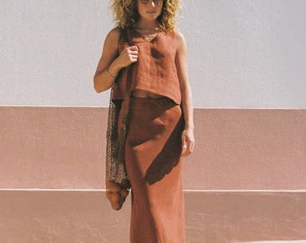 May include: A woman in a rust-coloured linen outfit, comprising a sleeveless top and a long skirt. She carries a mesh bag. The background features a white and pink wall and a concrete surface.