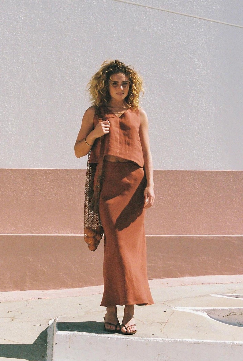 May include: A woman wearing a rust-colored linen outfit, consisting of a sleeveless top and a long skirt. She carries a mesh bag over her shoulder. The background features a white and pink wall and a concrete surface.