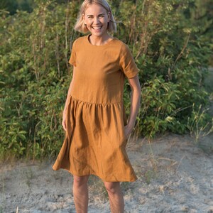 May include: A woman wearing a short-sleeved, rust-colored linen dress with a gathered skirt. She is standing in a field with green foliage in the background.