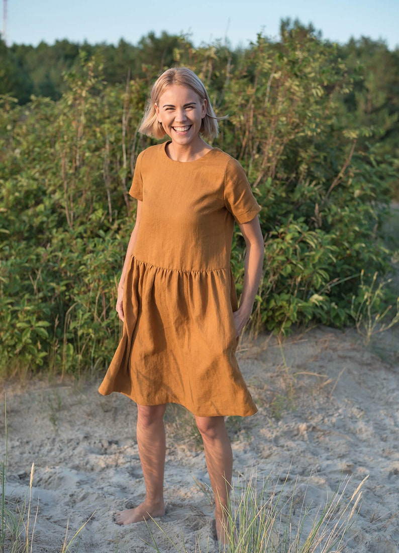 May include: A woman wearing a short-sleeved, rust-colored linen dress with a gathered skirt. She is standing in a field with green foliage in the background.