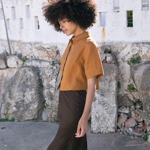 May include: A short-sleeved, button-down shirt in a warm, golden-brown hue paired with a long, dark brown skirt. The outfit is made of a lightweight fabric, creating a relaxed and comfortable look. The model is walking on a stone path.