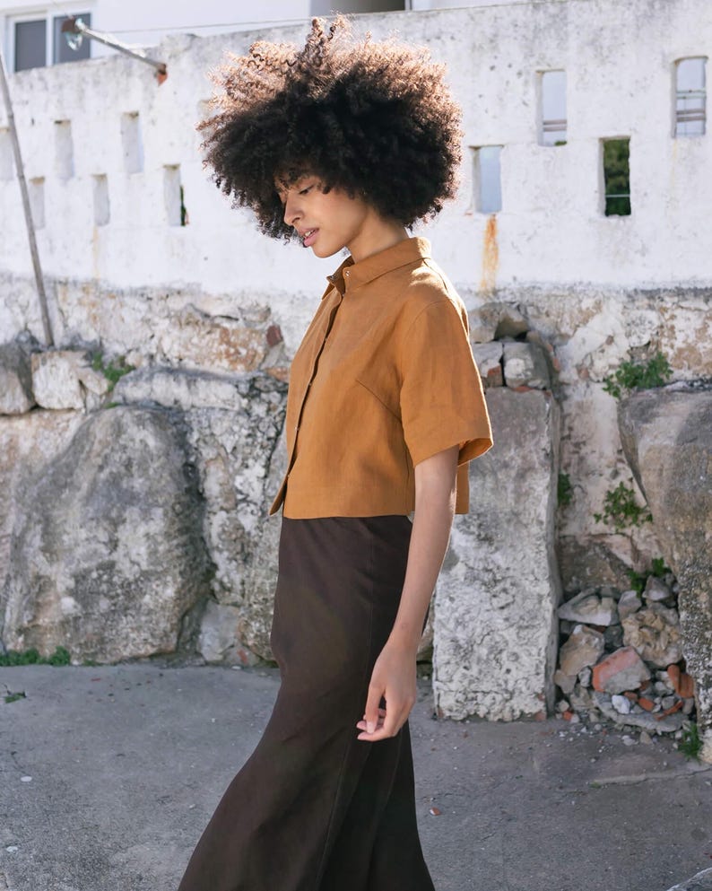 May include: A short-sleeved, button-down shirt in a warm, golden-brown hue paired with a long, dark brown skirt. The outfit is made of a lightweight fabric, creating a relaxed and comfortable look. The model is walking on a stone path.