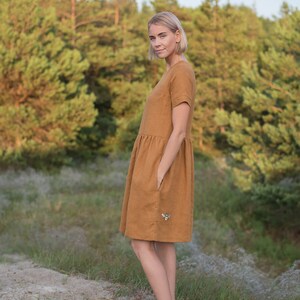 May include: A woman wearing a brown linen dress with a bee embroidered on the pocket. She is standing in a grassy area with trees in the background.