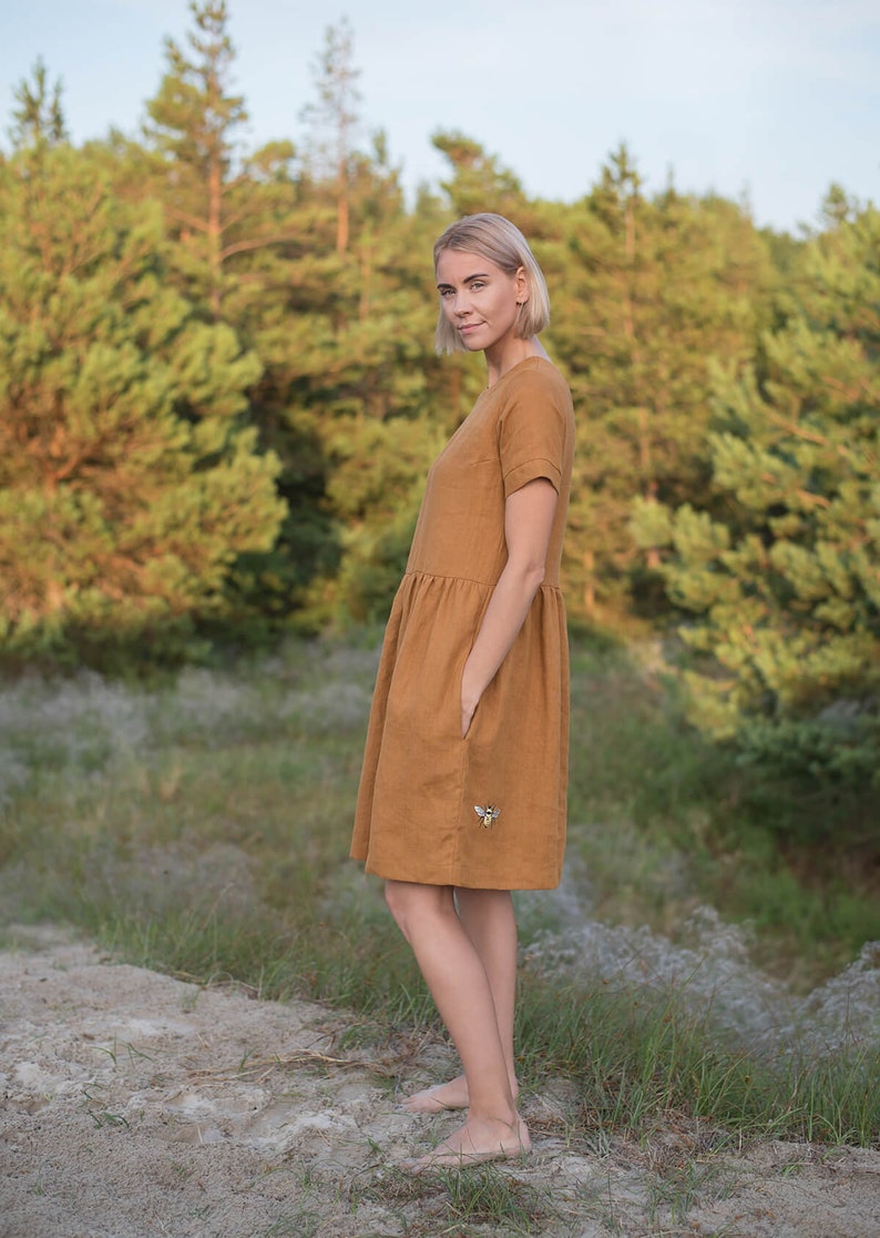 May include: A woman wearing a brown linen dress with a bee embroidered on the pocket. She is standing in a grassy area with trees in the background.